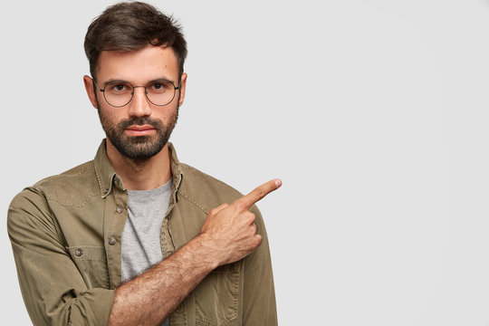 Horizontal shot of handsome unshaven young male with dark stubble, strict look, has serious expression, dressed in fashionable shirt, points right aside, shows free space for advertising content
