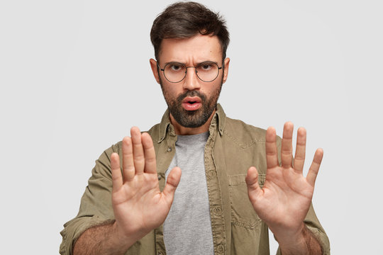 Photo of bearded young male shows stop gesture, has displeased facial expression, denies something, talks about forbidden things, wears fashionable shirt, isolated over white studio background