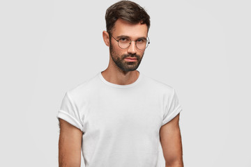 Waist up portrait of handsome bearded male with serious expression, contemplates about something, wears round spectacles and white casual t shirt, poses indoor. People and facial expressions