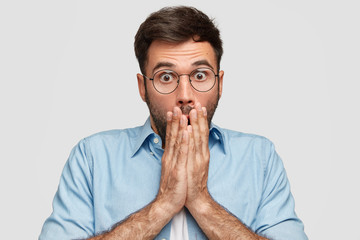 Puzzled Caucasian man covers mouth with both hands, has scared puzzled expression, can`t believe in failure, stares through eyewear with bugged eyes, dressed in elegant shirt, stands indoor.