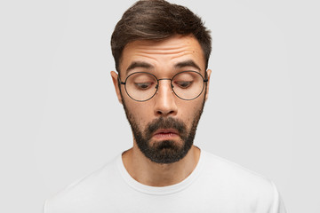Curious unshaven male worker focused down, looks with interest as notices something, wonders, being in white t shirt and round eyewear, poses alone against white background. Facial expressions - obrazy, fototapety, plakaty