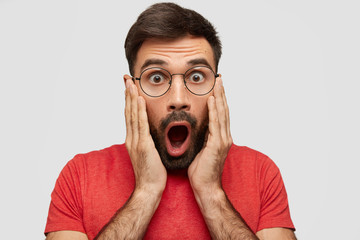Horizontal portrait of astonished bearded male touches cheeks with hands, stares with bugged eyes,...
