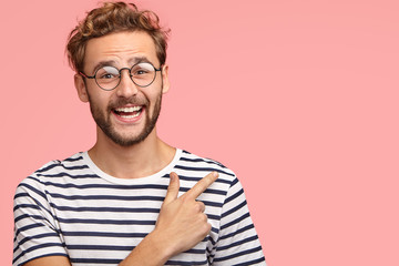 Attractive unshaven man with curly hairstyle, looks positively at camera as indicates at blank space for advertising content, isolated over pink studio wall. Hey, look here! Cheerful hipster indoor