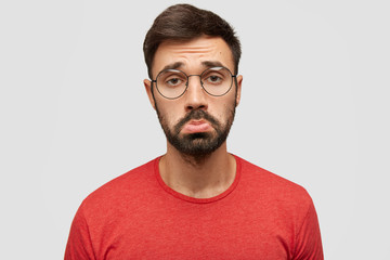 Fototapeta premium Gloomy displeased sorrowful bearded young male purses lips in discontent, being offended by bad comments of followers, expresses negativity, wears red jacket, stands against white studio wall