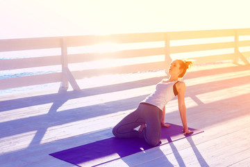 Young woman practicing yoga outdoors at white wooden seafront. Sitting in easy pose and bending...