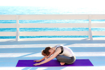 Young woman practicing yoga outdoors at white wooden seafront. Sitting in easy pose and bending...