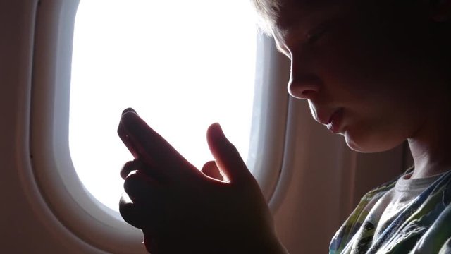 Closeup view of cute profile of young kid sitting in economy class seat in plane near illuminator using smartphone to play video games during flight. Real time full hd video footage.