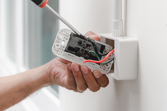 The electrician is using a screwdriver to attach the power cord to the wall outlet.