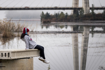 Niña escuchando música sentada al lado del río junto al puente de la ciudad