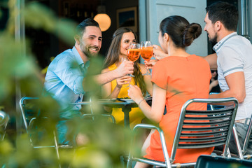 Cheerful young best friends toasting with a refreshing delicious summer drink while sitting together at table at a trendy restaurant outdoors