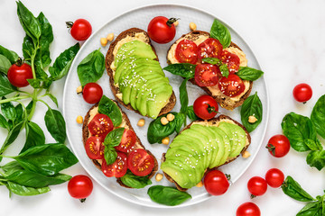 Avocado and tomatoes toasts with hummus, sesame and basil in a plate over white marble background....
