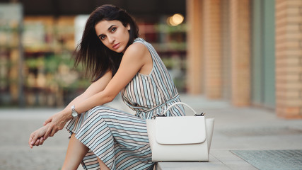 Woman fashion model posing sitting with bag in city