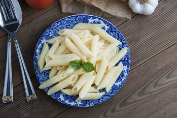 Cooked penne pasta with butter, cheese and fresh vegetable ingredients on background: tomato, garlic, onion, fork spoon and basil. Vintage beautiful blue plate on wooden table.