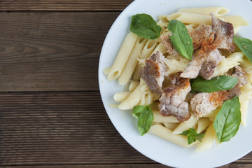 Cooked penne pasta isolated in white round plate, with basil leaves and pork meat, over wooden table, decorated with basil leaves. Top view.