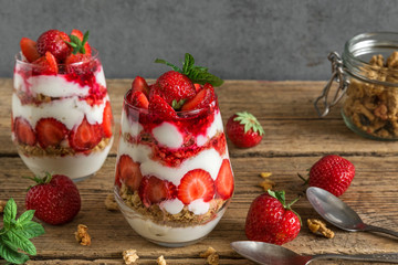 strawberry yogurt parfait with granola, mint and fresh berries in glasses with spoons on rustic wooden table