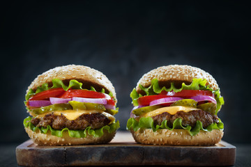 Two home made tasty grilled burgers with lettuce, cheese, onion and tomato on a rustic wooden plank on dark background