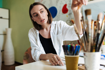 Young thoughtful girl at the desk dreamily choosing paint brush in workplace at home