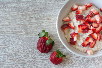 Top view of rolled  organic porridge with cut strawberries and two whole berries in marble bowl half is shown on wooden table  healthy breackfast
