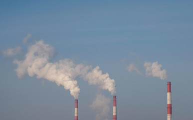 Three red and white pipes of thermal power station with smoke in sky and tree background.