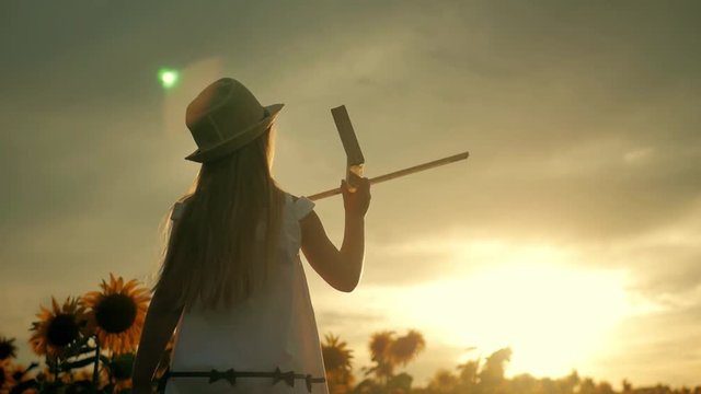 A girl with hat playing with a wooden airplane. Happy child playing with toy airplane on sunflower field on sunset. In the rays of the sun, the rear view. Baby dream, childhood, memories concept.