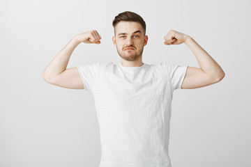 Strong good-looking male with beard and stylish hairstyle in white casual shirt raising hands showing mascules proudly being in great physical condition working out in gym bregging about biceps