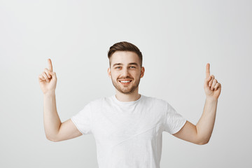 Pleasant friendly and handsome young caucasian male with brown stylish hairstyle raising hands and pointing up with broad satisfied smile offering drink coffee in cafe upstairs over gray bacground