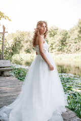 Fototapeta na wymiar A cute curly woman in a white wedding dress with a wedding bouquet and wreath in her hair standing back to the camera in nature. Concept escaped bride. Forward to a happy bright future Runaway