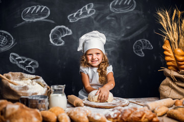 Charming little girl with curly hair in white apron and hat standing at table kneading bread dough...
