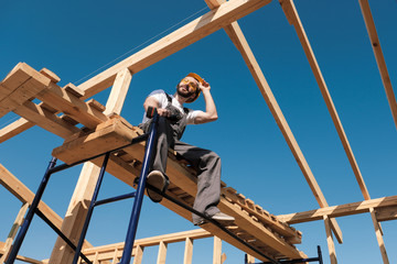 The man builder sits on the edge of the roof of the frame house, in a yellow helmet and gray overalls. The blue sky and clear sunny day.