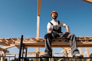 The man builder sits on the edge of the roof of the frame house, in a yellow helmet and gray...
