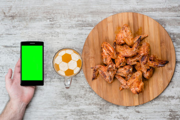Male hand holding smartphone mobile cell phone near mug with a football on a beer foam and grilled chicken wings, food delivery order app application concept
