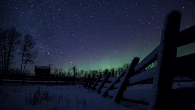 Time lapse of stars, milky way and northern lights over a snow covered wooden fence at a deserted farm in rural Alberta, Canada