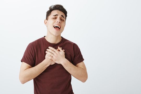 Indoor shot of passionate good-looking boyfriend in trendy red t-shirt, holding palms on heart and singing serenade with closed eyes, confessing in love and tender feeling over gray background
