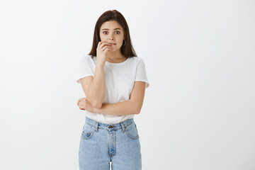 Studio shot of worried impatient cute caucasian girl in stylish street style outfit, biting fingernail and gazing at camera while waiting for results nervously, standing anxious over gray background