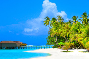 Water villas in turquoise ocean and wooden walkway leading to the island with white beach and palm trees