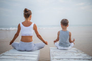 Fototapeta premium A mother and a son are doing yoga exercises at the seashore of tropic ocean