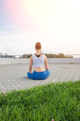 Fototapeta na wymiar Young pretty woman practices yoga and meditates on the sunny embankment of the river