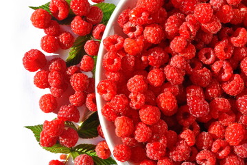 Lots of harvested red ripe juicy sweet fresh raspberry in a large plate and next on green leaves. White background. Isolated. Top veiw