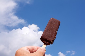 hand holding a Popsicle on a stick against a blue sky with clouds