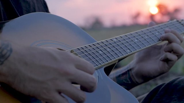 Young tattooed male play on acoustic guitar in summer fields during beautiful sunset