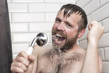 Cute bearded man singing in the bathroom using the shower head with flowing water instead of a...