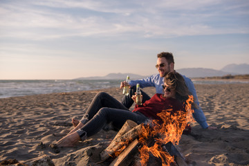 Young Couple Sitting On The Beach beside Campfire drinking beer