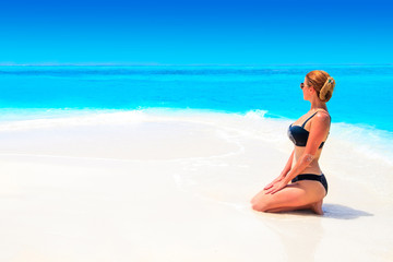 Blond woman with black bikini tans on the sandbank in front of turquoise ocean and blue sky