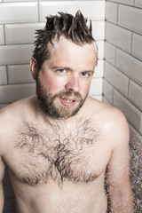 Handsome, bearded man in a white foam from the shampoo standing in the shower room.