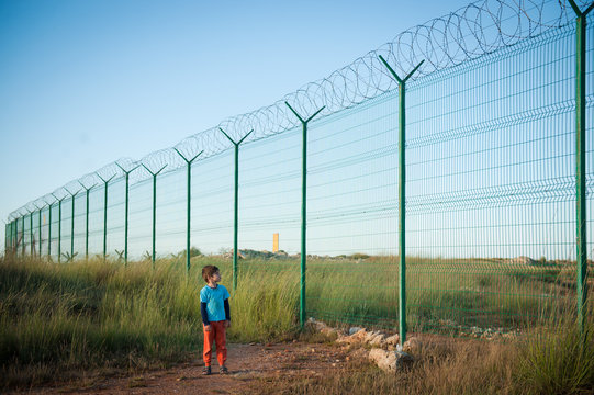 lonely little kid refugee watching at high fence with barbed razor wire desert