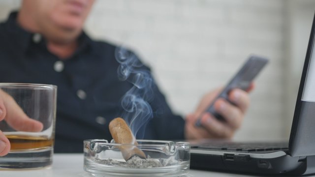 Blurred Image with a Businessman Smoking Cigar Drinking Whisky and Using Cellpho