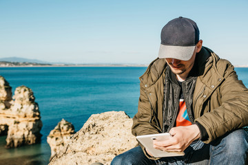A young man or a tourist uses a tablet sitting on the Atlantic coast next to the city called Lagos in Portugal. Use of mobile applications or maps or freelancing or remote work or communication.