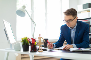 Fototapeta premium A young man sits at a table in the office, holds a red pen in his hand and works with documents.