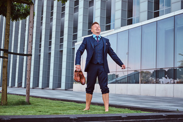 Confident businessman dressed in elegant suit holds his shoes enjoying while standing barefoot on a green lawn against cityscape background.