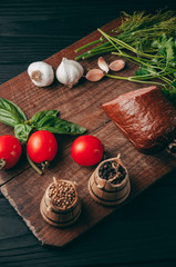 Smoked salami on a black table and a brown board. Sausages with rosemary, garlic and pepper and tomato cherry, dill, parsley.
